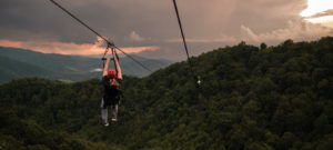 person ziplining across forested mountain below with sunset in background